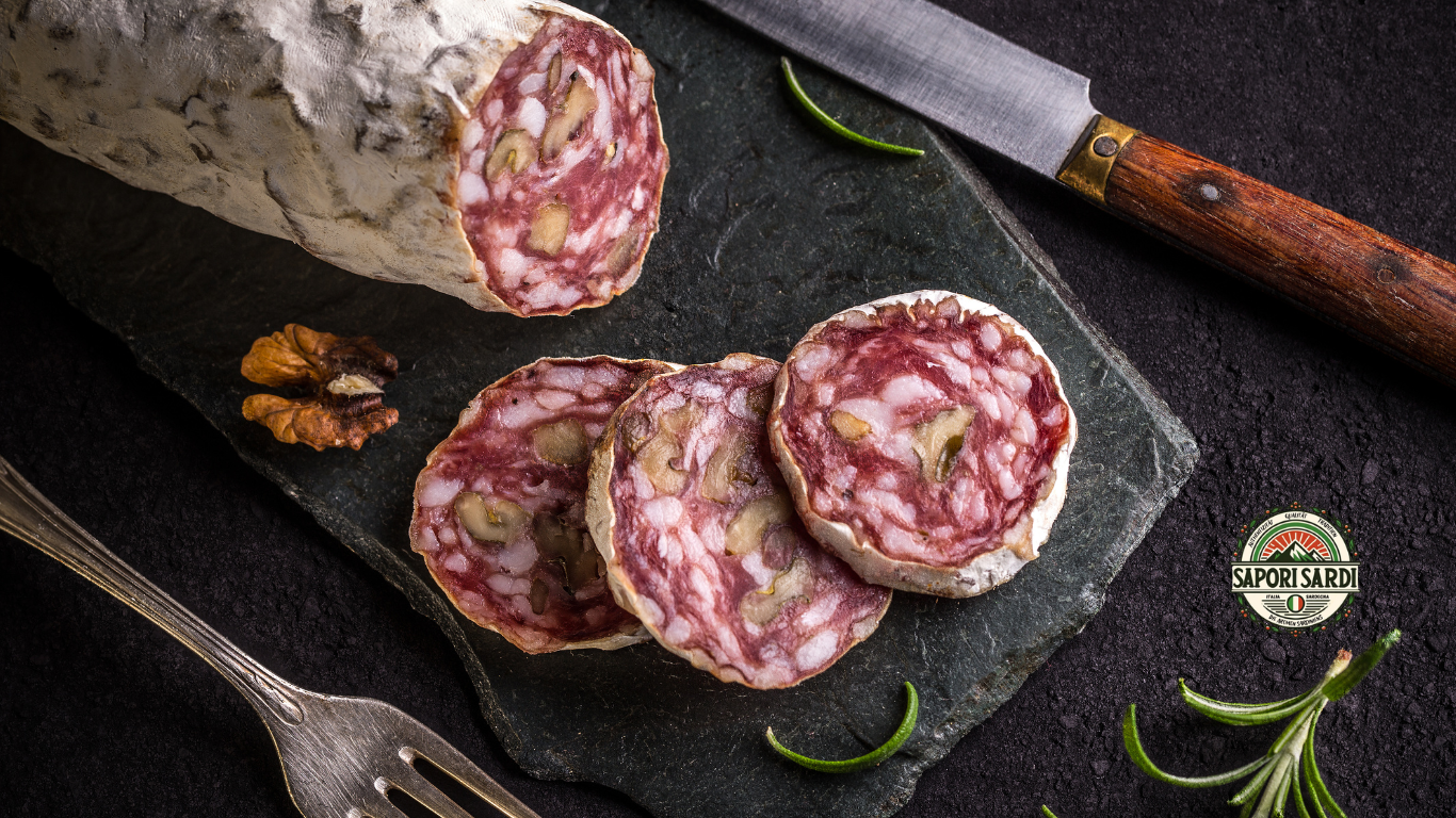 Aufgeschnittene Salami auf einer Schiefertafel. Oben sieht man ein altes Messer mit Holzgriff. Unten rechts ist das Logo von Sapori Sardi zu sehen. Rosmarin und eine halbe Wallnuss verzieren das Brett.