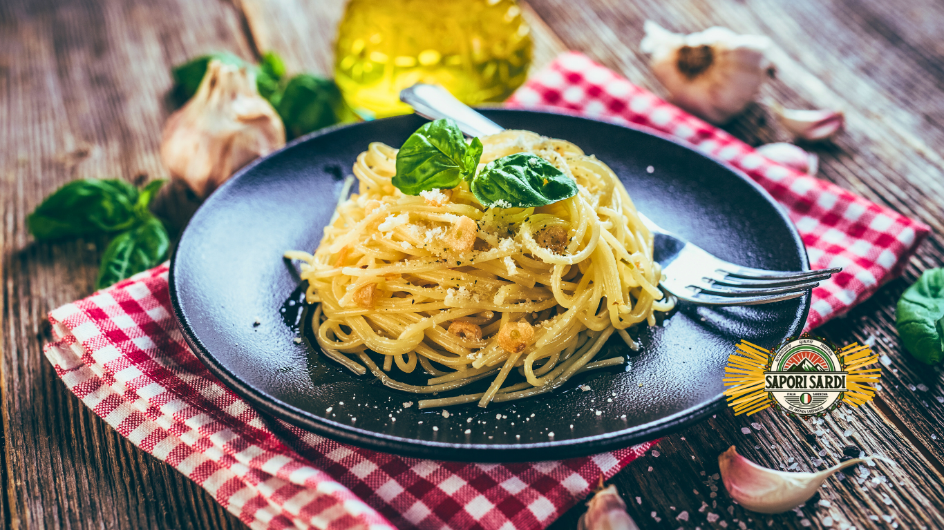 Spaghetti aglio e olio auf einem Teller schwarzen angerichtet. Der Teller steht auf einem Holztisch mit einer rot weiß karierten Serviette darunter. Als Dekoration findet man Knoblauch, Basilikum und Olivenöl auf dem Tisch.