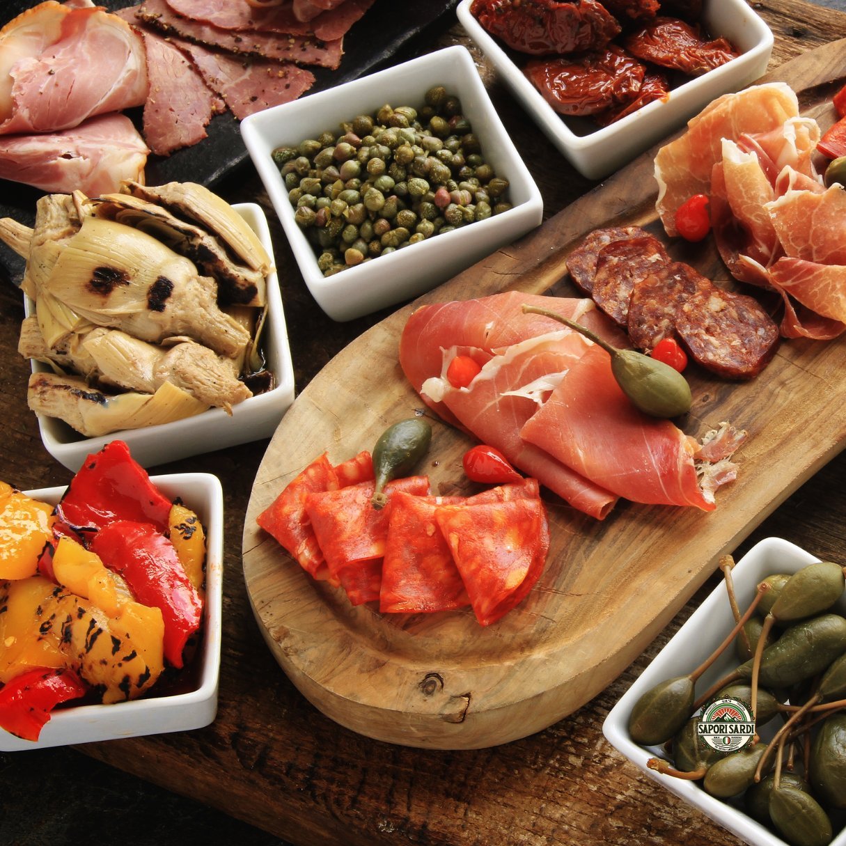 Verschiedene Antipasti aus Sardinien in Schalen und auf einem Holzbrett angerichtet. Darunter Gebratene Artischocken, gebratene Paprika, Salami und Schinken.