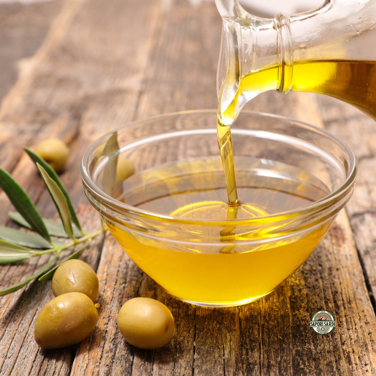 Extra Vergine Olivenöl aus Sardinien welches aus einer kleinen Karaffe in eine kleine Schale gefüllt wird. Die Schale steht auf einem Holztisch und ist umgeben von ein paar frischen Oliven und Olivenblättern als Dekoelemente.