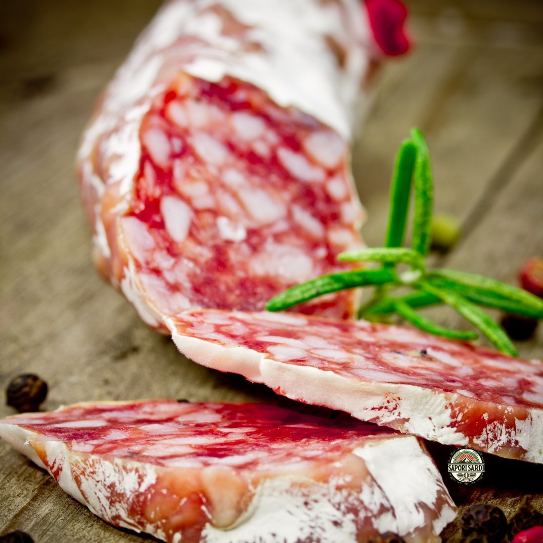 Frisch aufgeschnittene Salami aus Sardinien. Diese Salsiccia ist eine Delikatesse. Sie ist mit etwas Rosmarin verziert. und liegt auf einem Holzbrett. Pfefferkörner liegen dekorativ um die Salami herum.