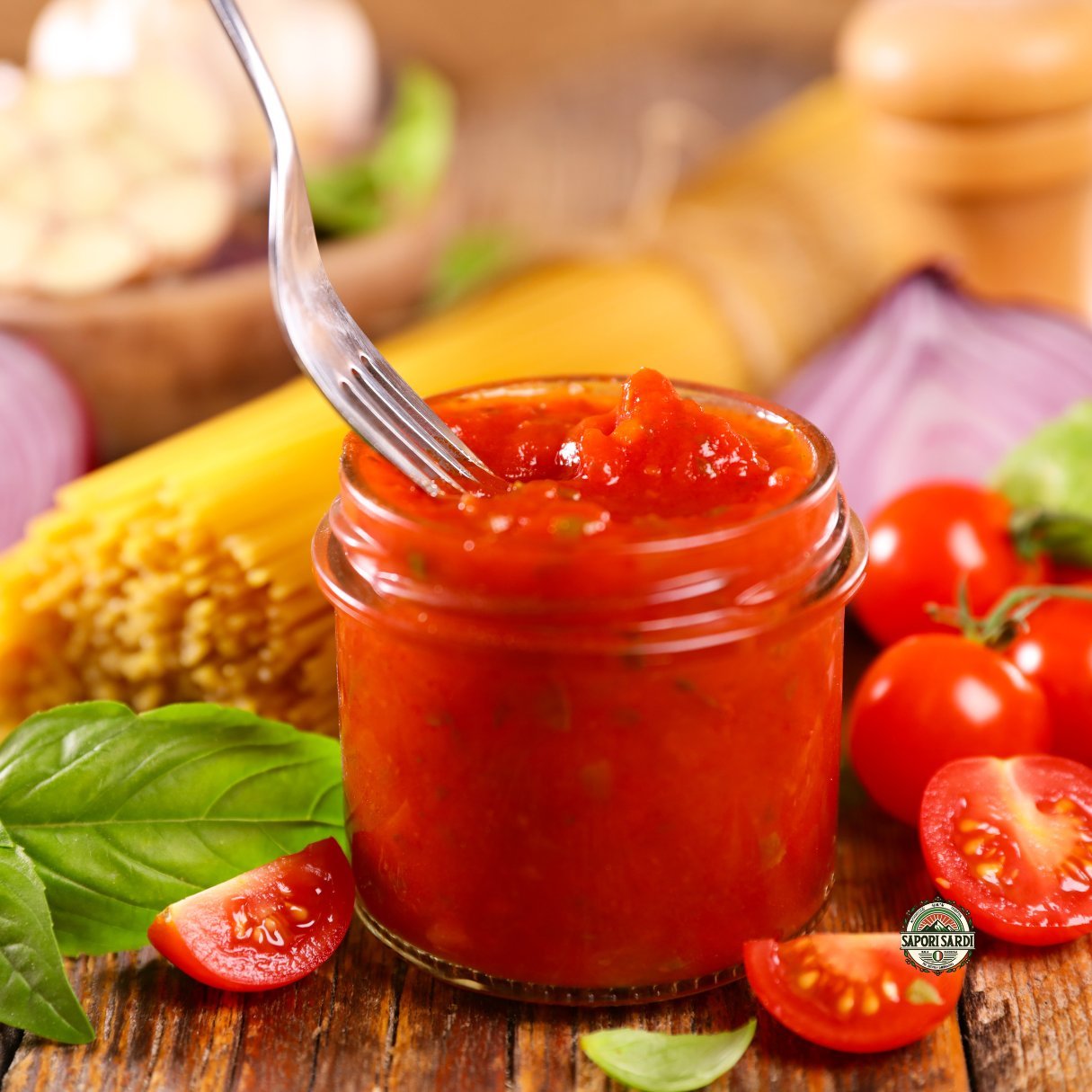 Frische Tomatensoße, welche gerade in ein kleines Glas gefüllt wurde. Im Hintergrund sieht man Pasta und der Holztisch auf dem das Glas steht ist mit Tomatenstücken und Basilikum verziert.