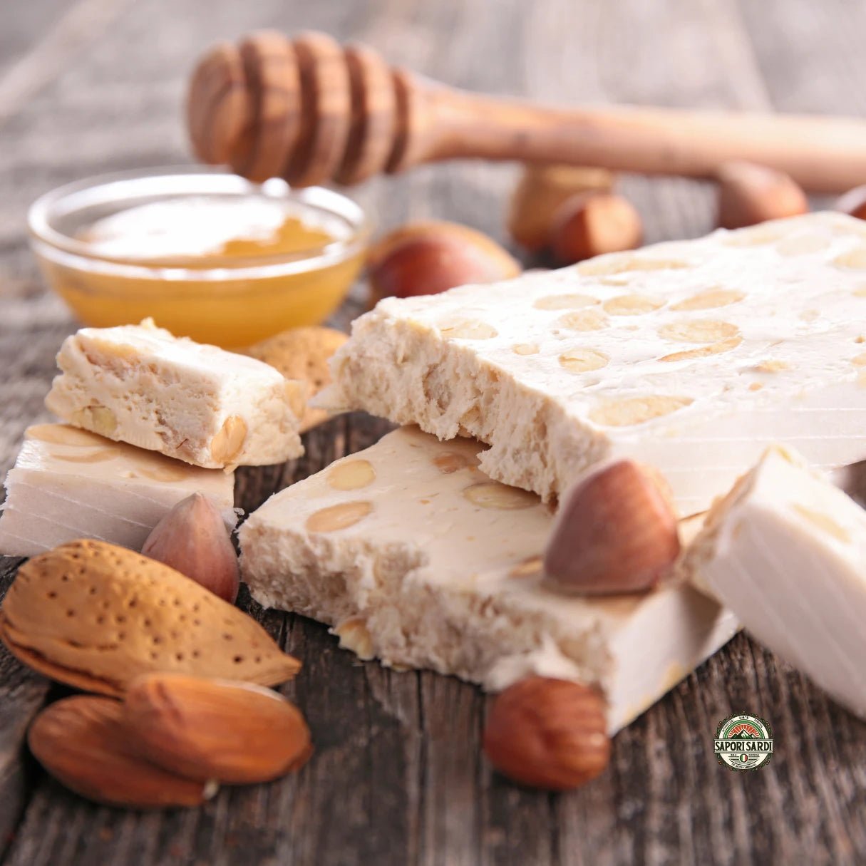 Frischer Torrone aus Sardinien auf einem Holztisch. Er ist bereits etwas aufgebrochen und bereit zu Verzehr. Als Dekoelemente sieht man eine Schale Honig mit einem Honiglöffel und einige Nüsse.