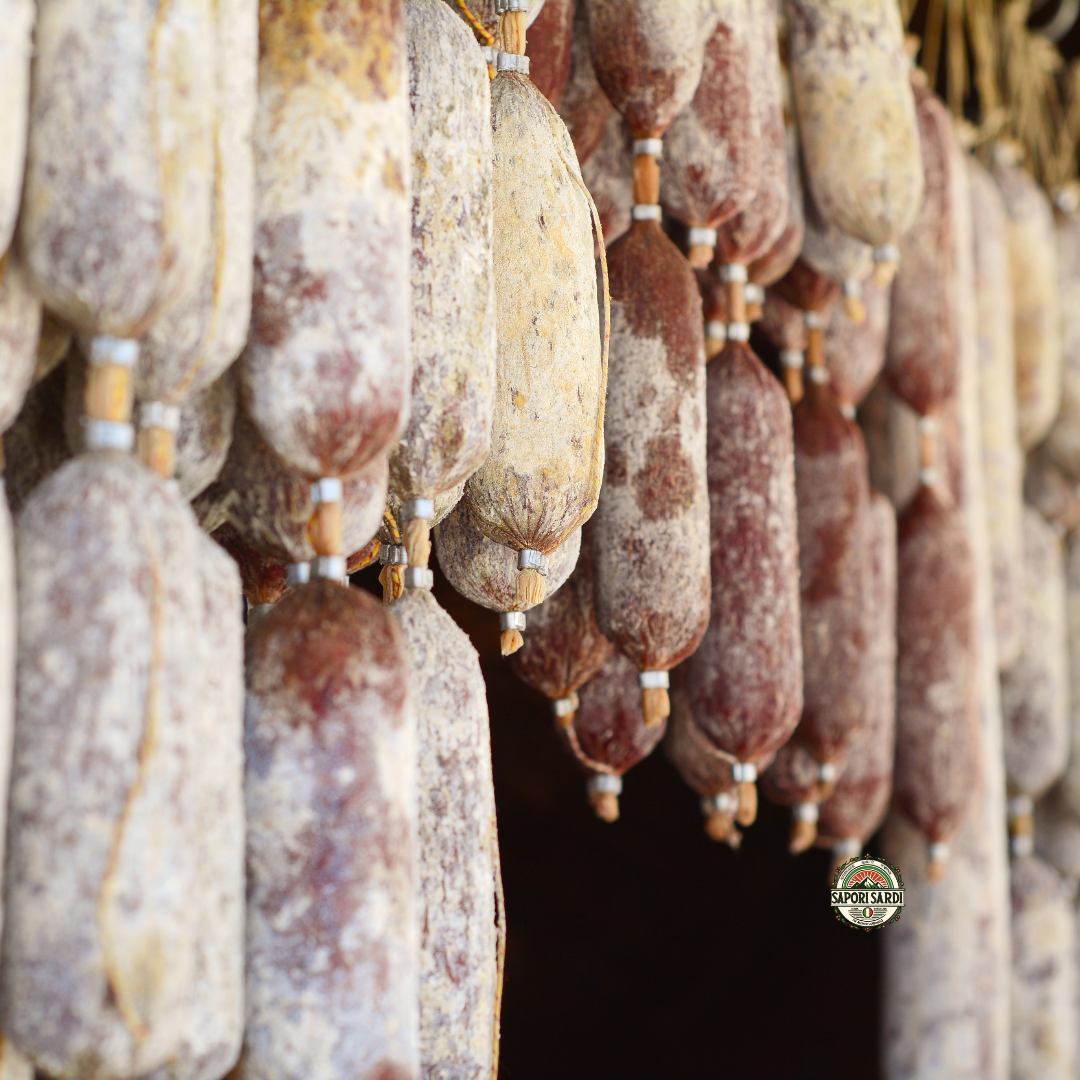 Salami aus Sardinien hängt zum Reifen in einem Keller nebeneinander aufgehangen. Unten ist das Logo von Sapori Sardi zu erkennen.