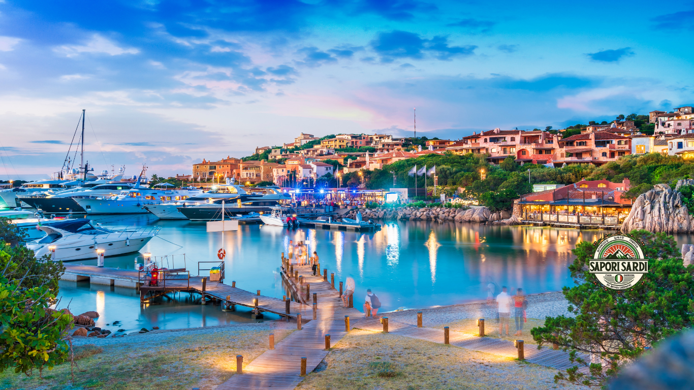 Ein Yachthafen in Olbia. Das Bild wurde vom Strand aufgenommen und zeigt große beleuchtete Yachten am Abend im Hafen stehen. Dahinter das Dorf. Unten rechts im Bild ist das Logo von Sapori Sardi.