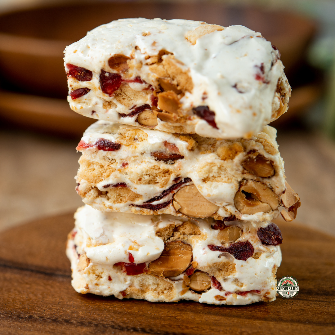Weißer Torrone Stücke gestapelt auf einem Holzbrett. Im Torrone sind Nüsse und Beeren zu sehen. Unten rechts ist das Logo von Sapori Sardi zu sehen.