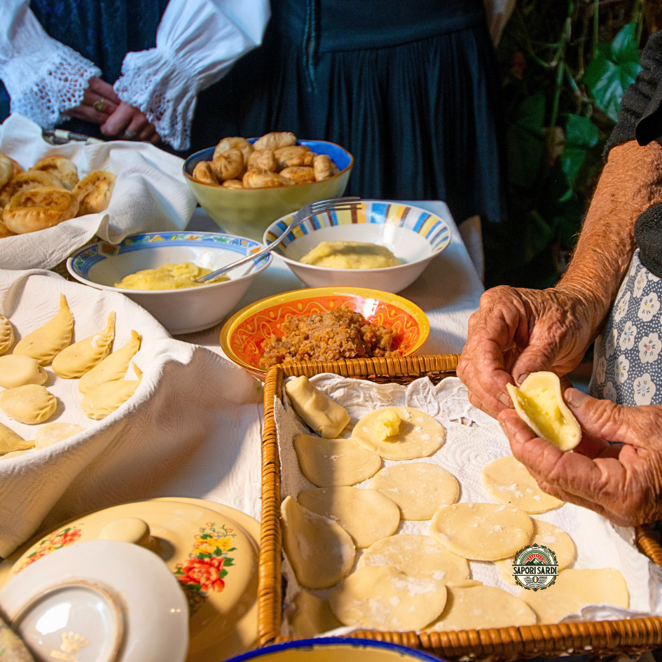Culurgiones werden traditionell per Hand gefaltet. Man sieht mehrere Schalen mit Zutaten und einen Korb mit kreisförmigem Teig aus welchem eine Dame die Culurgiones formt.