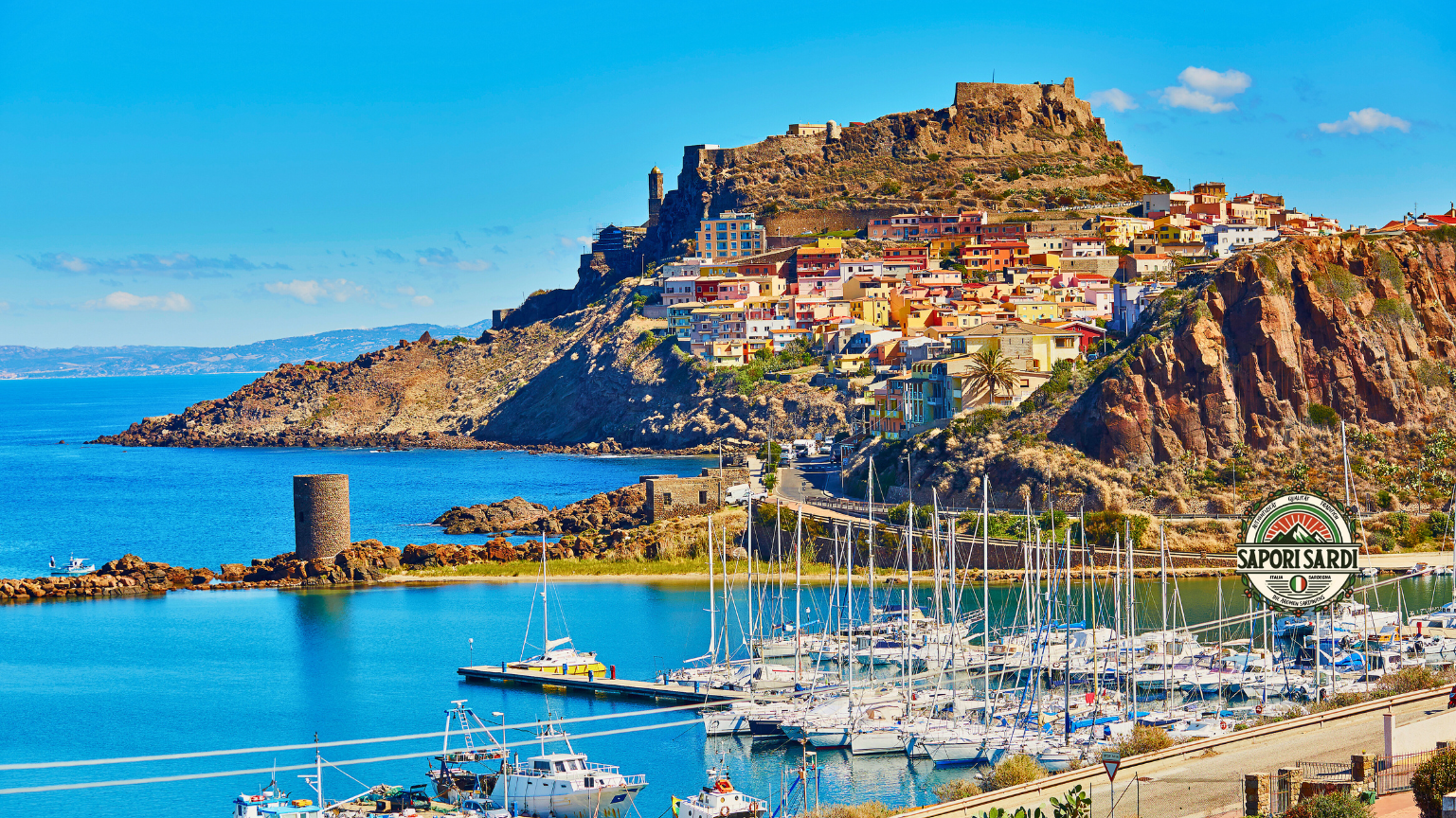 Castelsardo auf Sardinien. Das Bild wurde von einer Erhöhung aufgenommen und zeigt die mittelalterliche Stadt aus der Ferne. Im Hintergrund das Meer und vor der Stadt der Hafen mit Segelbooten. Unten rechts im Bild ist das Logo von Sapori Sardi.