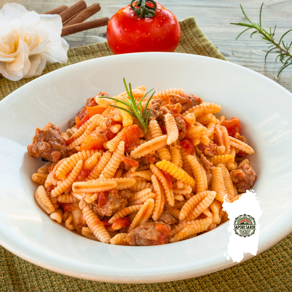 Malloreddus alla Campidanese auf einem weißen Teller angerichtet. Der Tisch ist verziert mit einer Tomate, Zimtstangen und Rosmarinzweigen. Unten rechts im Bild ist das Logo von Sapori Sardi zu erkennen, welches auf einer weißen Karte von Sardinien liegt.